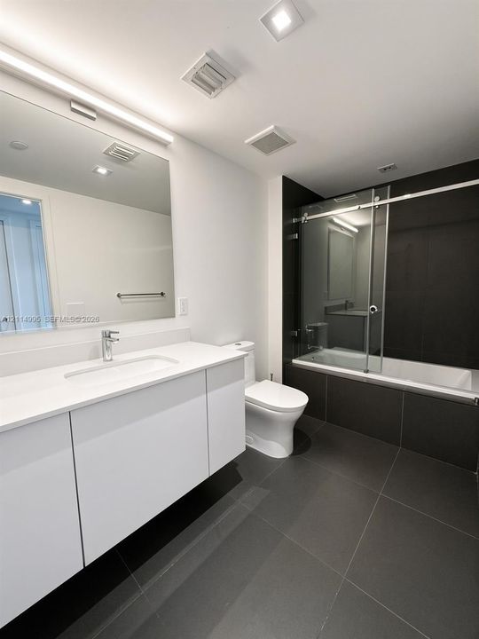 Modern floating vanity with a solid-surface countertop and integrated sink, complemented by a large wall-mounted mirror, sliding glass shower door, and large-format tile flooring