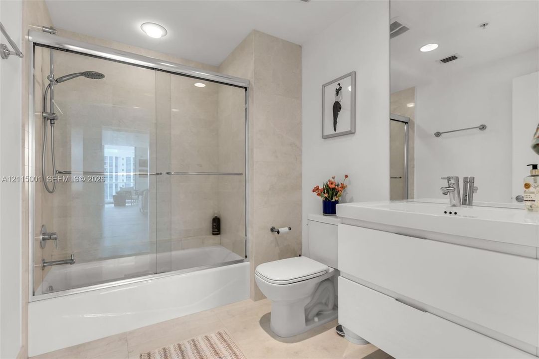 Bathroom featuring a white floating dual-drawer vanity with integrated sink, chrome faucet, wall-spanning mirror, and a tub-shower combination enclosed by a sliding glass door with stone-finish wall tile