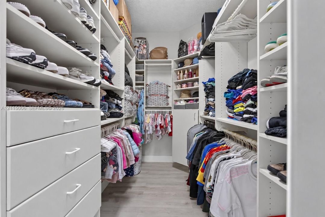Custom walk-in closet featuring white built-in shelving, multiple hanging rods, integrated storage drawers with bar pulls, and wood-finish flooring