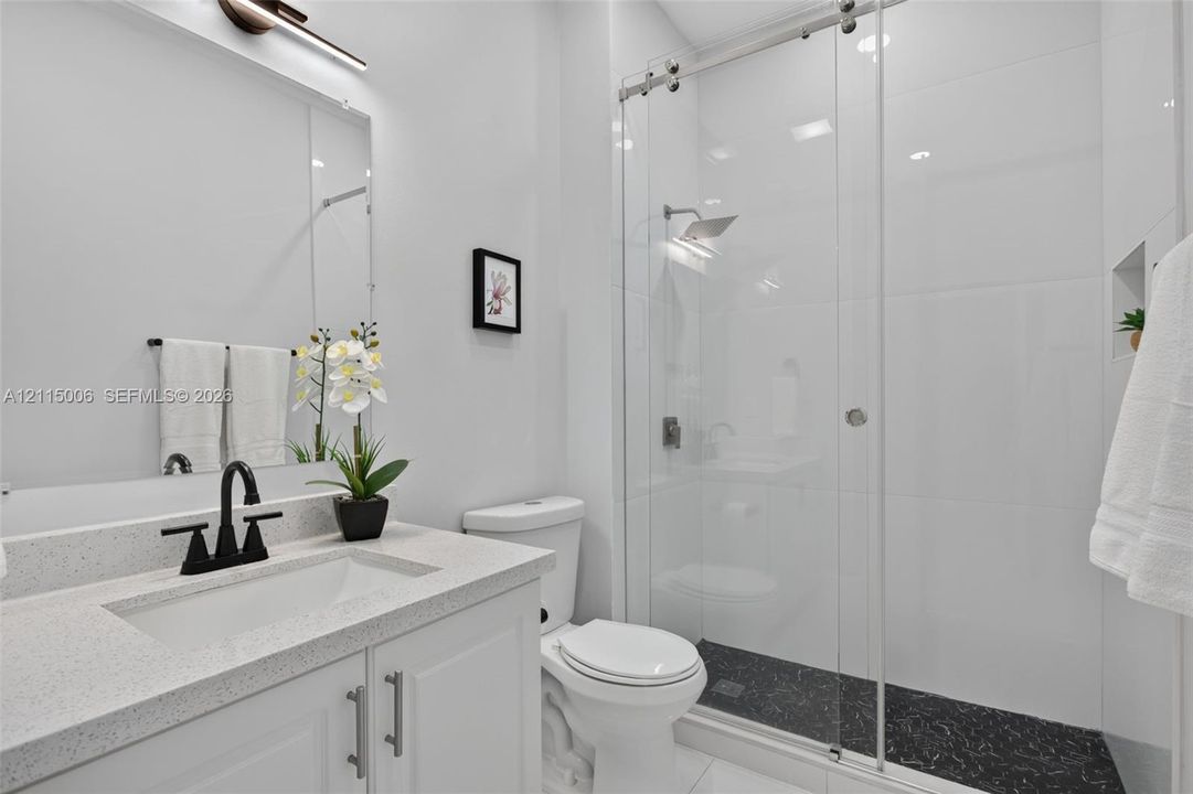White cabinetry vanity with solid-surface countertop, matte black faucet, glass-enclosed shower with contrasting dark stone-finish flooring, and wall-mounted light fixture