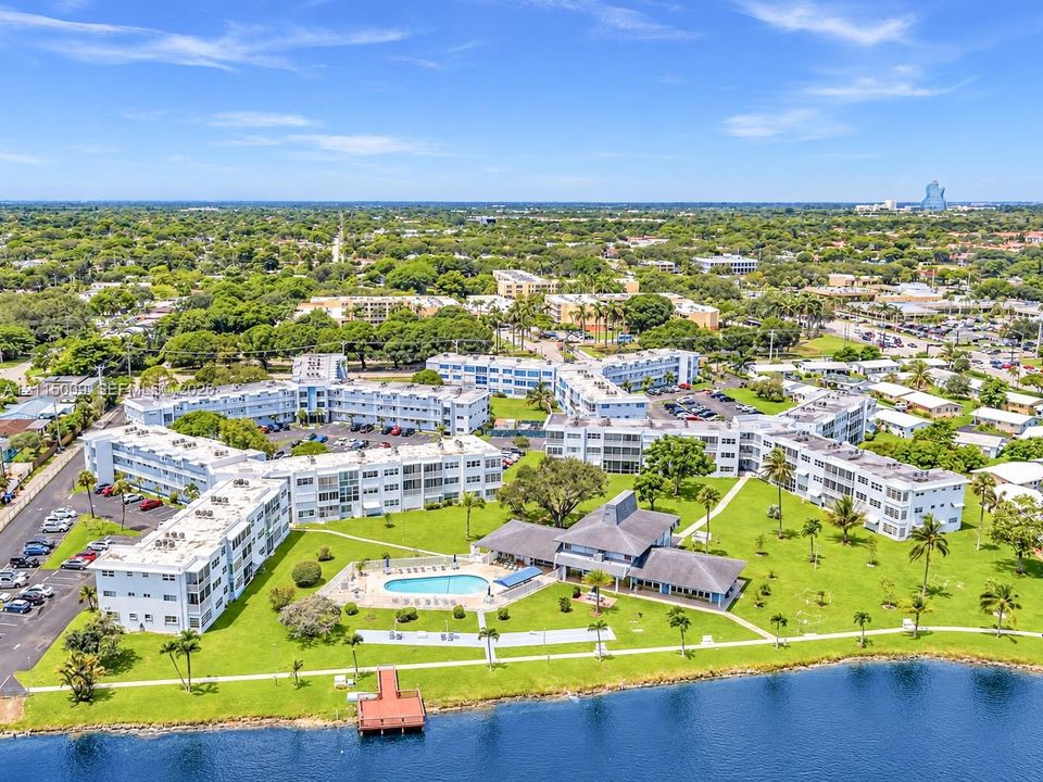 Aerial perspective of a multi-building residential complex featuring a community swimming pool, waterfront dock, expansive green space, and paved surface parking