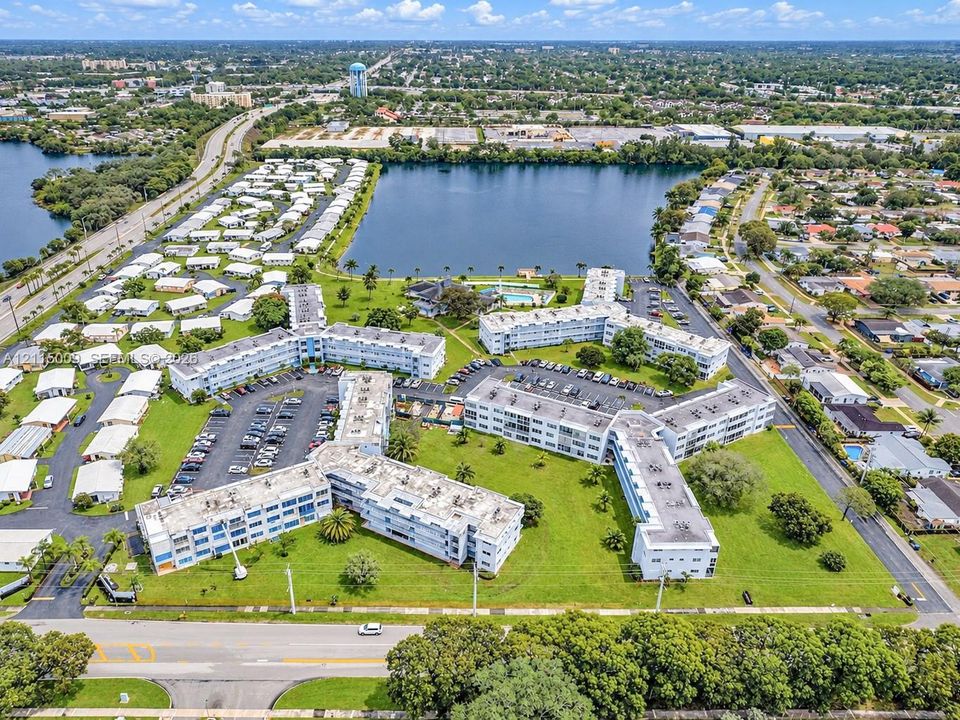 Aerial perspective of a multi-story residential complex featuring waterfront positioning, expansive landscaped grounds, surface parking, and a dedicated swimming pool area
