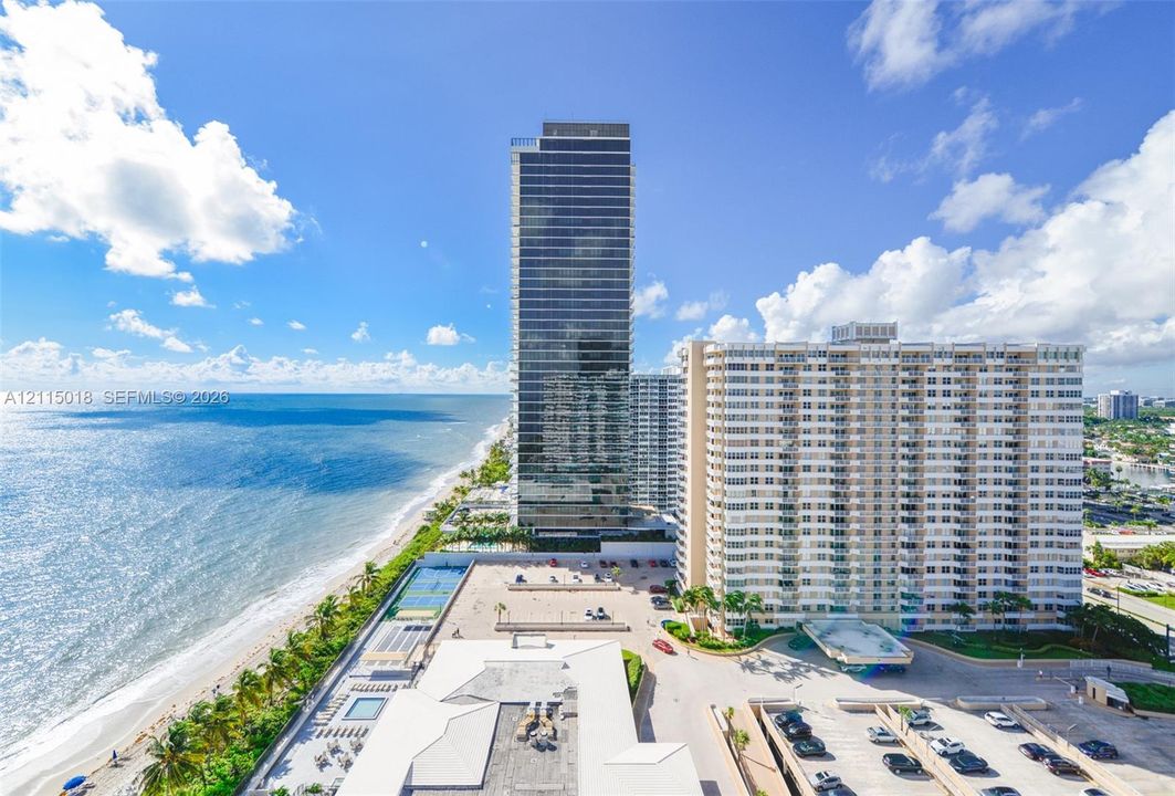 High-rise residential tower exterior featuring floor-to-ceiling windows, direct oceanfront positioning, integrated ground-level tennis courts, and expansive structured parking facilities