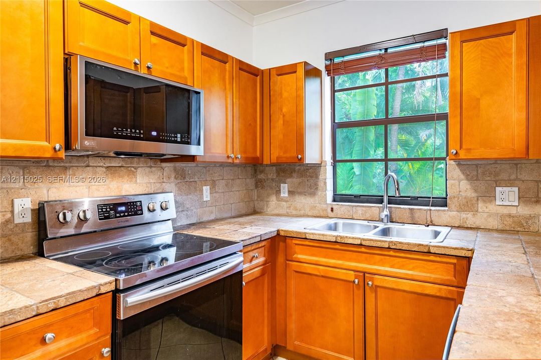 Kitchen featuring wood cabinetry, stainless steel appliances, natural stone tile countertops, and a stone tile backsplash with an integrated double-basin sink