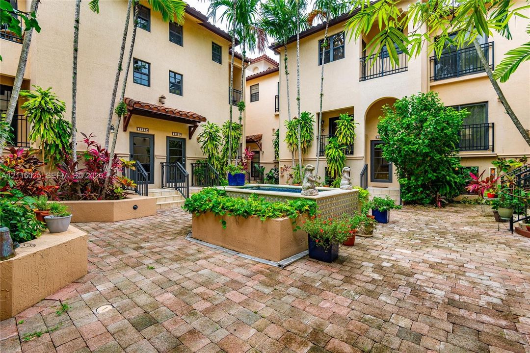 Stucco exterior featuring arched entryways, private balconies with black metal railings, a central fountain with tile detailing, and paver-stone courtyard flooring