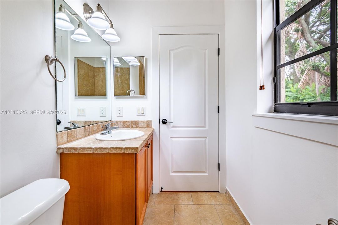 Bathroom featuring a wood-finish vanity with a tiled countertop, oversized wall mirror, three-light fixture, and natural stone-finish floor tiling