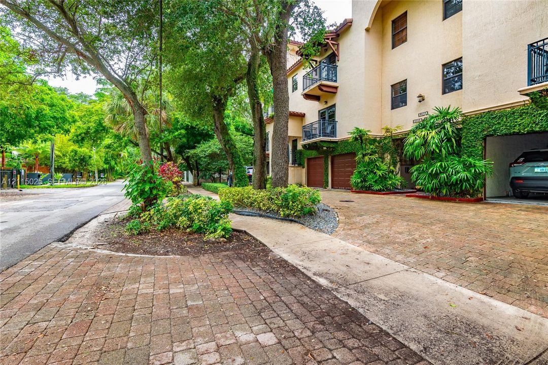 Multi-story exterior featuring a textured stucco facade, integrated covered parking, private balconies with dark metal railings, and brick-paver hardscaping surrounded by mature landscaping