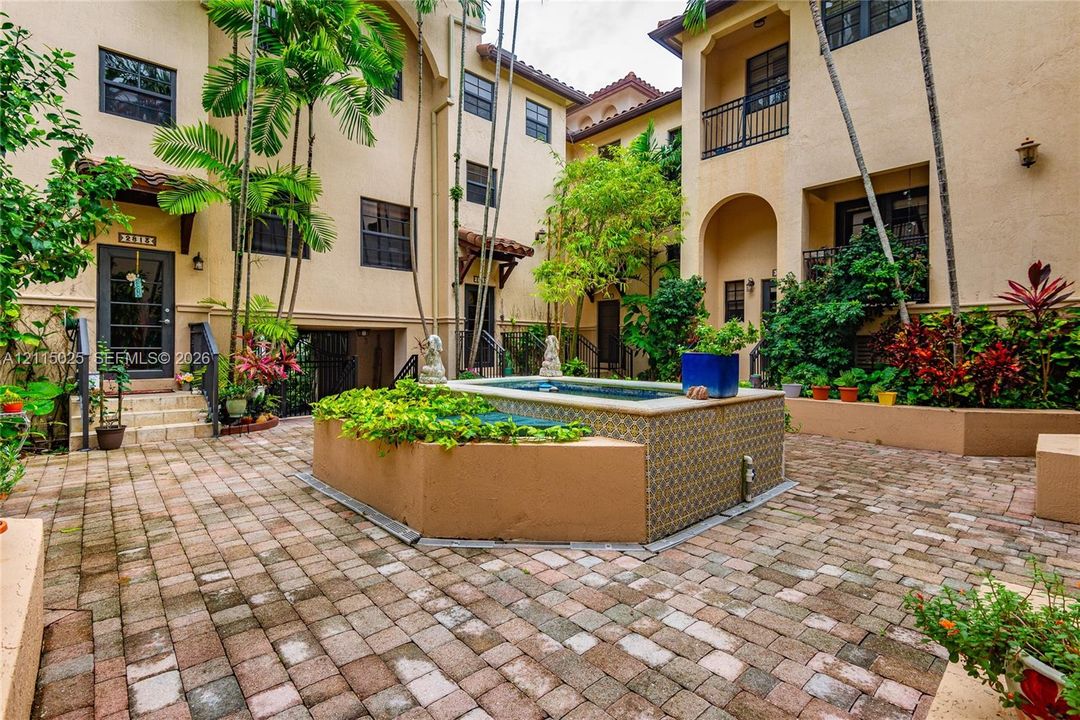 Mediterranean-style courtyard featuring a paved brick patio, elevated decorative tile-accented water feature, arched architectural detailing, and wrought-iron balcony railings