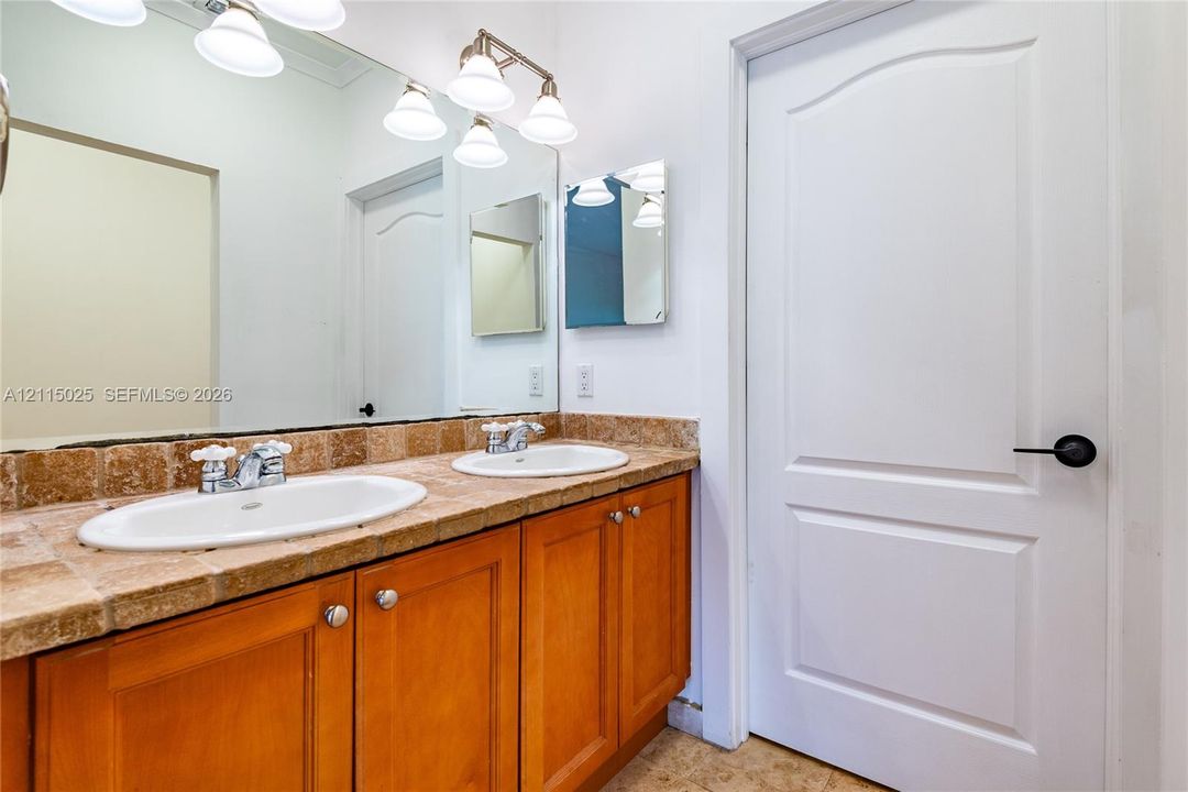 Dual-sink vanity with tile countertop, wood cabinetry, expansive wall-mounted mirror, and multi-bulb overhead lighting fixture