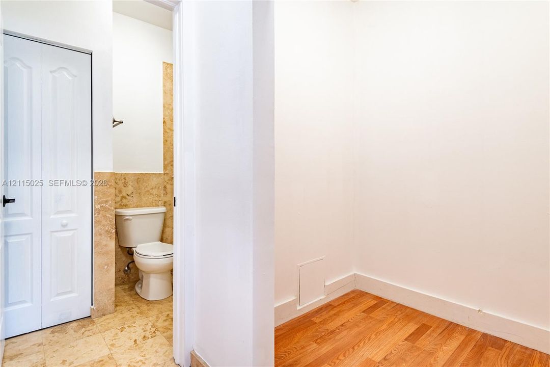 Storage area featuring wood-finish flooring and white bi-fold closet doors, with an entryway to a restroom finished with natural stone wall and floor tile