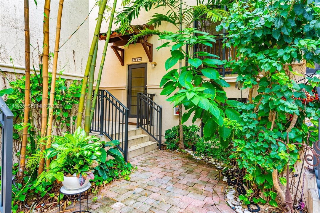 Entryway featuring brick-paver hardscaping, metal railings, a wooden door canopy, and mature landscaping