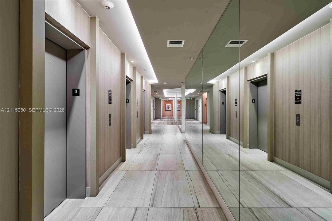 Hallway featuring wood-finish wall paneling, large-format tile flooring, a floor-to-ceiling mirrored wall, recessed linear lighting, and integrated elevator access