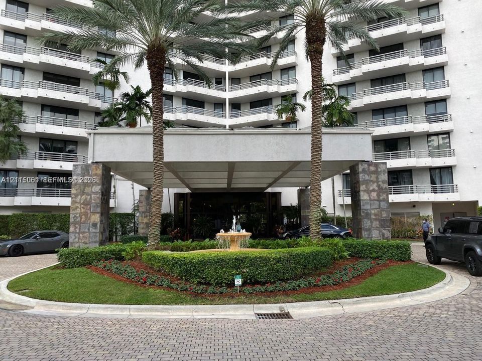 Multi-story residential building exterior featuring a covered porte-cochère, stone-clad structural pillars, a central fountain, and manicured circular landscaping