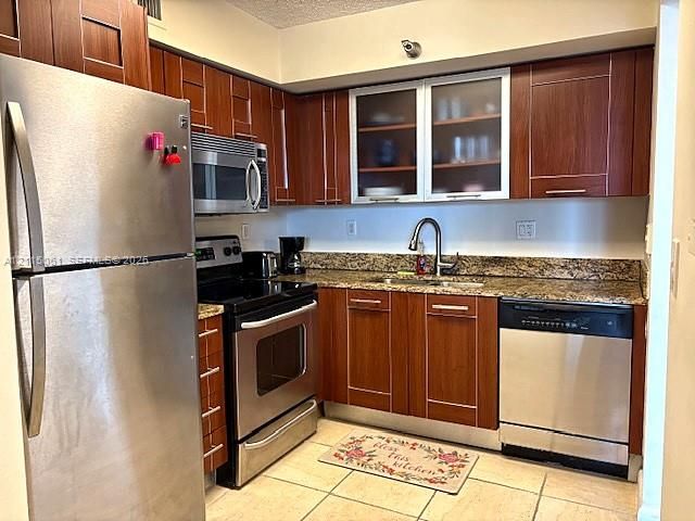 Kitchen featuring flat-panel wood-finish cabinetry, stone countertops, stainless steel appliances, glass-front upper cabinet doors, and tile flooring