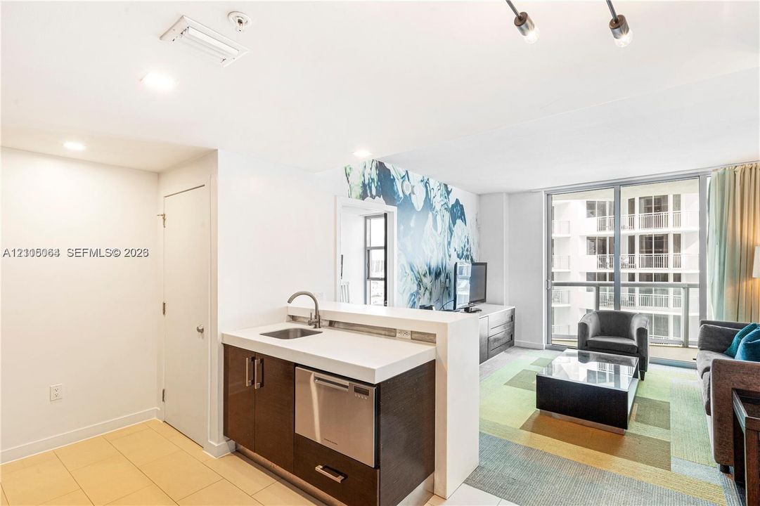 Kitchenette featuring dark cabinetry, solid-surface countertop, stainless steel undermount sink, and integrated stainless steel dishwasher, complemented by floor-to-ceiling glass panel windows