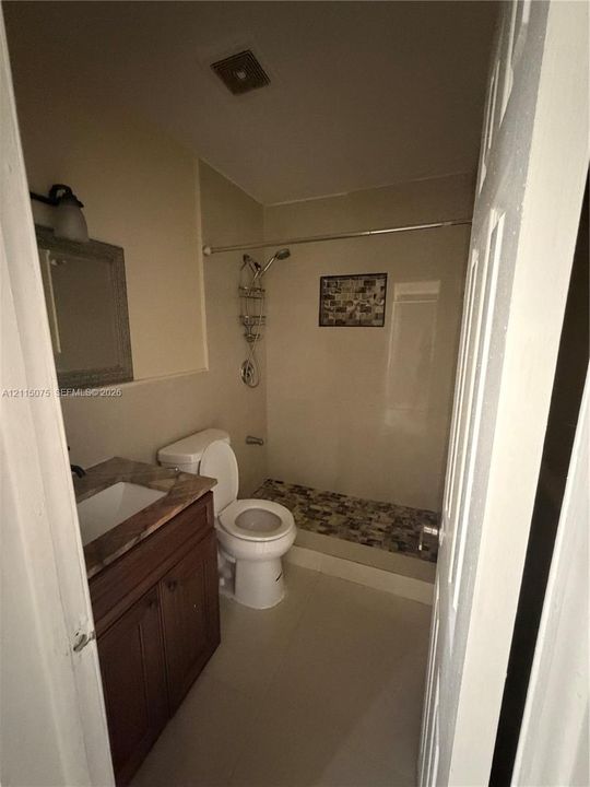 Bathroom featuring a wood-finish vanity with a stone-surface countertop, an integrated rectangular sink, a shower with a multi-textured stone-tile floor, and wall-mounted brushed metal fixtures