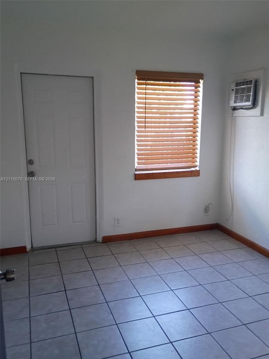 Light-filled room featuring neutral tile flooring, a window with wood-finish blinds, an exterior-access door, and a wall-mounted climate control unit