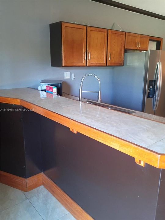 Kitchen peninsula featuring tiled countertop, wood-trim edge, stainless steel sink with high-arc pull-down faucet, and upper wood cabinetry