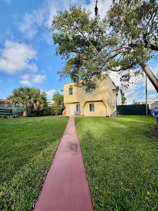 Two-story stucco exterior featuring a concrete walkway, expansive grass lawn, mature shade trees, and perimeter fencing