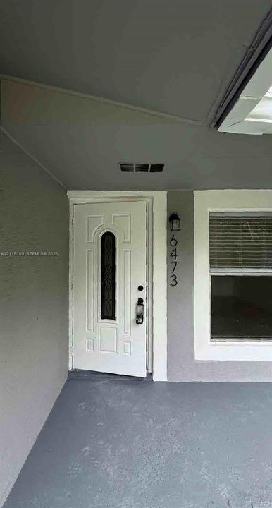 Covered porch entry featuring a white paneled door with an elongated decorative glass insert, wall-mounted light fixture, and neutral exterior finish