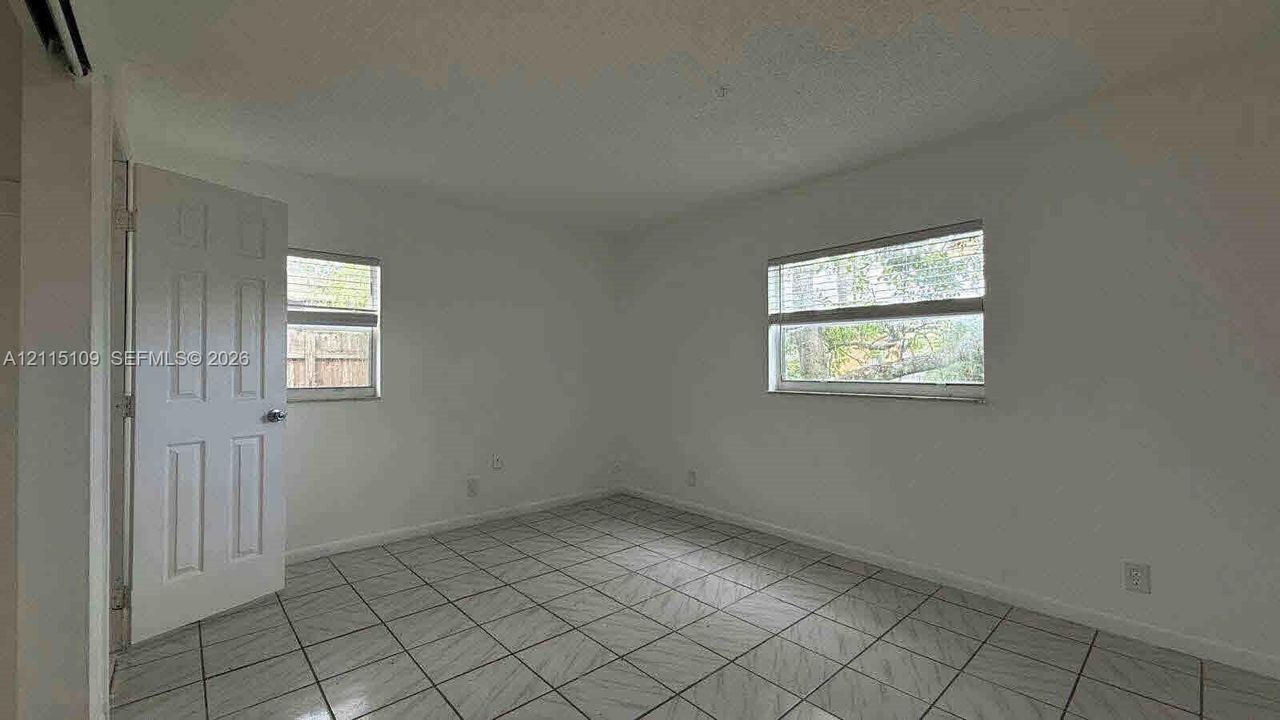 Neutral-toned interior space featuring grid-patterned floor tiling, smooth walls, two windows with horizontal blinds, and a hinged interior door