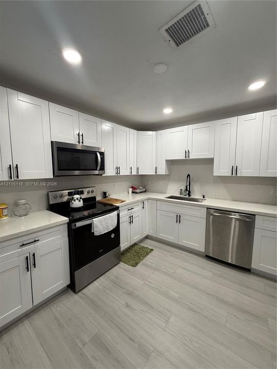 Kitchen featuring white Shaker-style cabinetry, stainless steel appliances, light-toned countertops, a stainless steel undermount sink with a matte black faucet, and wood-finish flooring
