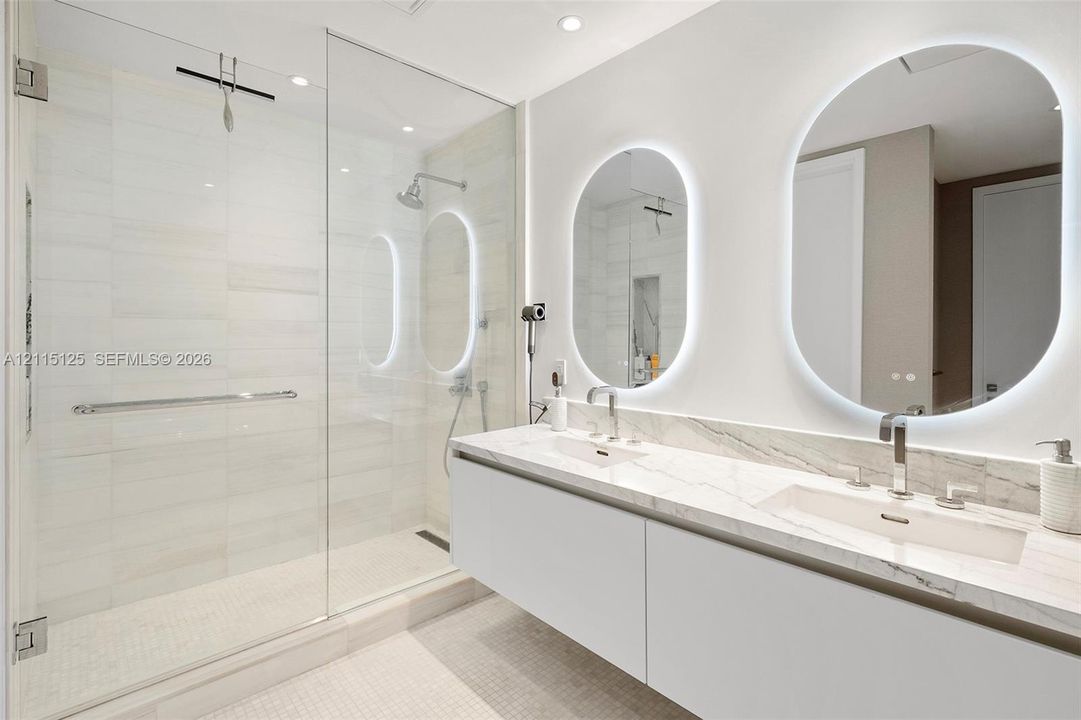 Floating dual vanity with stone countertop, integrated sinks, chrome fixtures, backlit oval mirrors, and a glass-enclosed shower featuring tile surrounds
