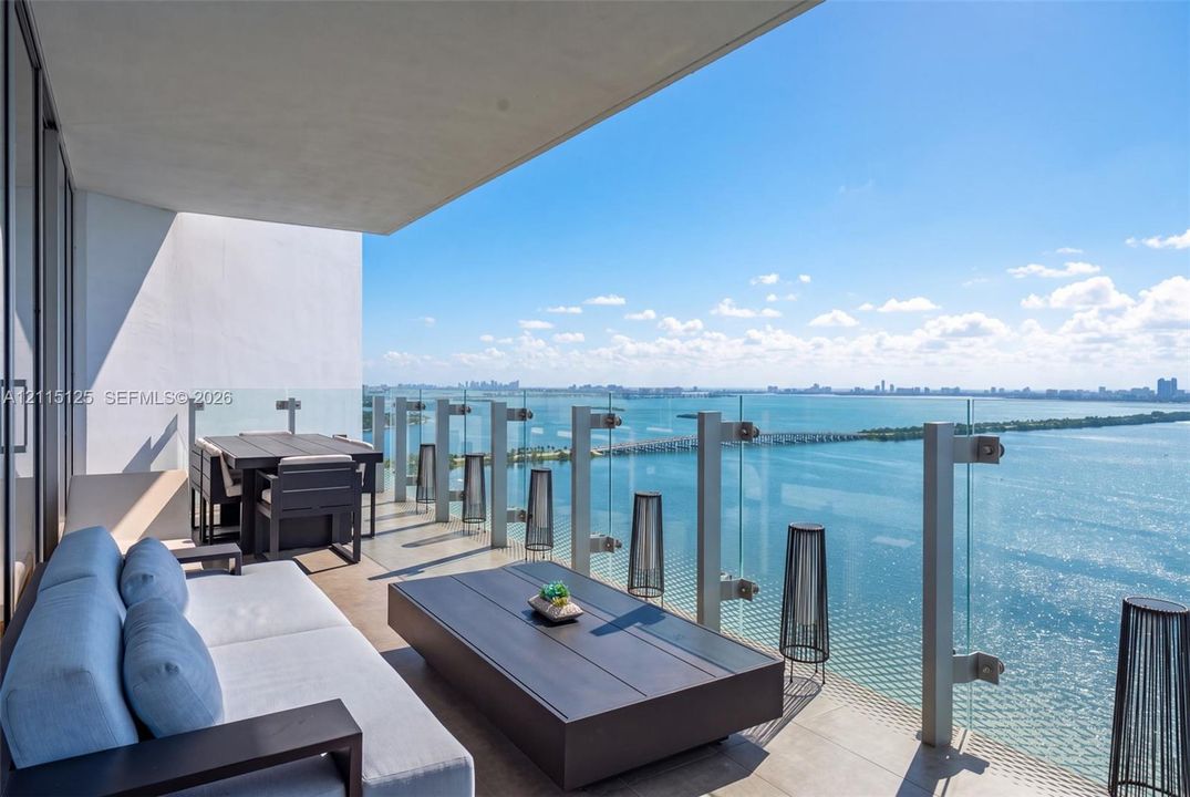 Floor-to-ceiling sliding glass doors, private balcony with glass railing, recessed tray ceiling with integrated cove lighting, matte black ceiling fan, and large-format tile flooring