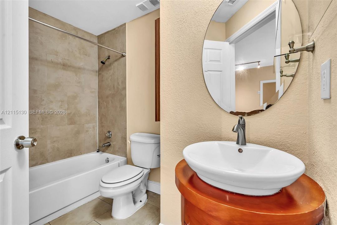 Oval wall-mounted mirror, vessel sink with brushed metal faucet, cylindrical wood-finish vanity, and shower-tub combination featuring tiled surround