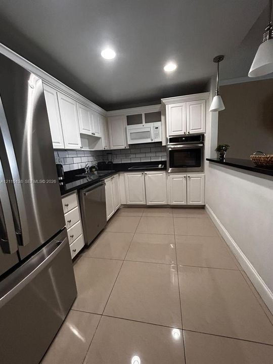 Kitchen featuring white shaker-style cabinetry, dark stone countertops, subway tile backsplash, stainless steel appliances including a built-in wall oven, and large-format tile flooring