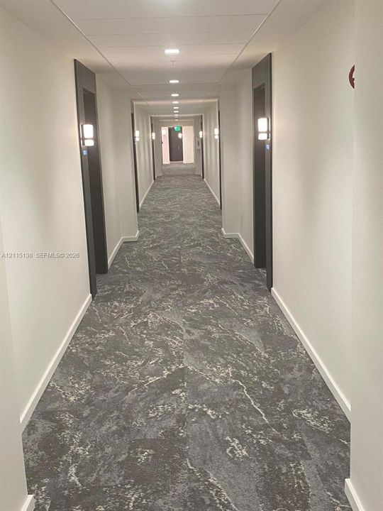 Hallway featuring patterned carpet flooring, recessed ceiling lighting, and wall-mounted light fixtures with dark door frames