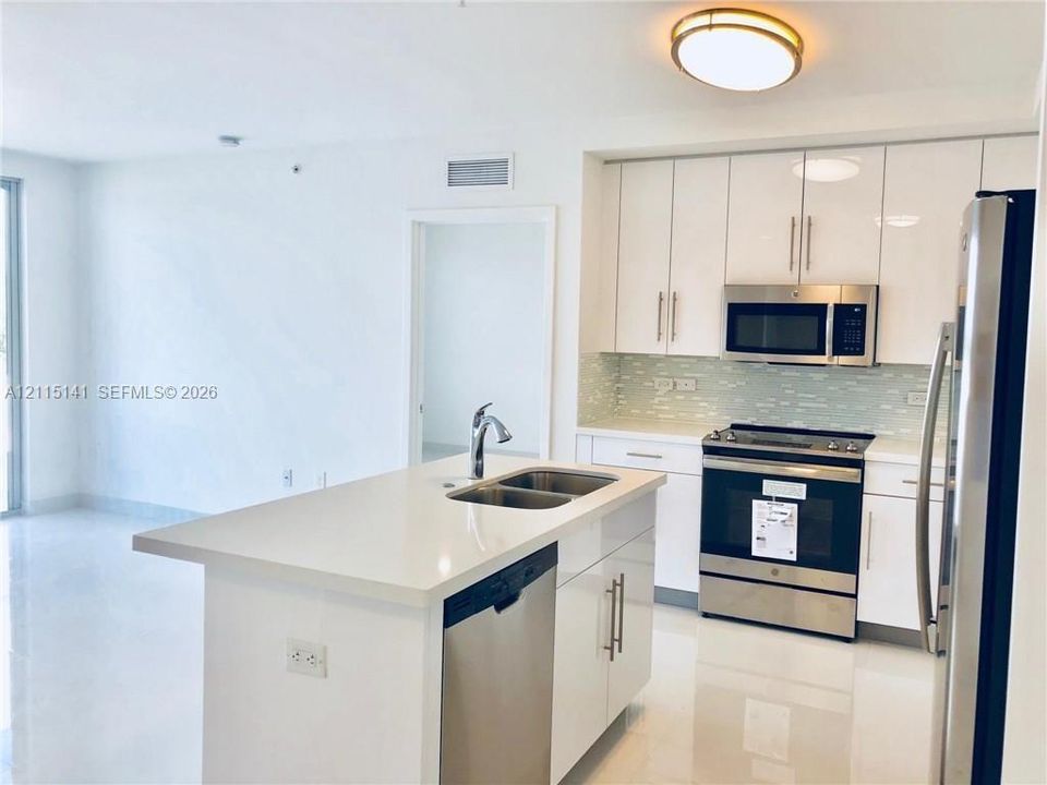 Kitchen featuring high-gloss white cabinetry, quartz countertops, stainless steel appliances, a central island with a dual-basin sink, and glass tile backsplash