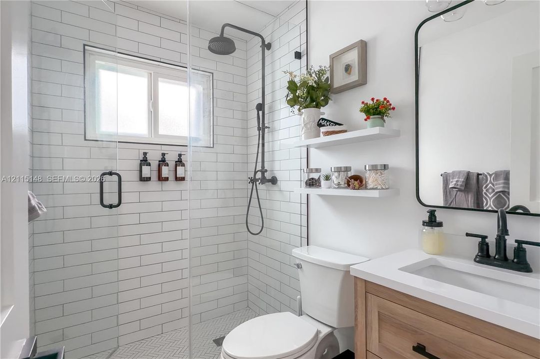 Walk-in shower with white subway tile surround, matte black rainfall showerhead, glass enclosure, wood-finish vanity with white countertop, and integrated wall-mounted shelving