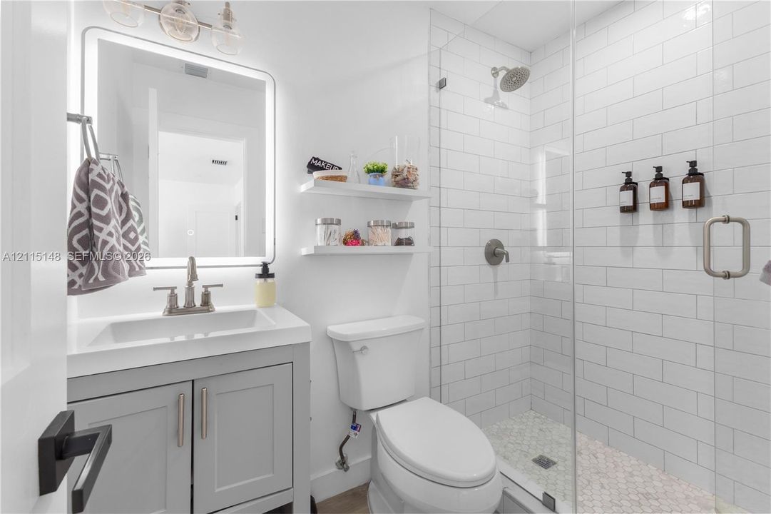 Bathroom featuring a gray shaker-style vanity with a rectangular sink, wall-mounted floating shelves, an illuminated vanity mirror, and a walk-in shower with white subway tile surround and pebble-style floor tiling