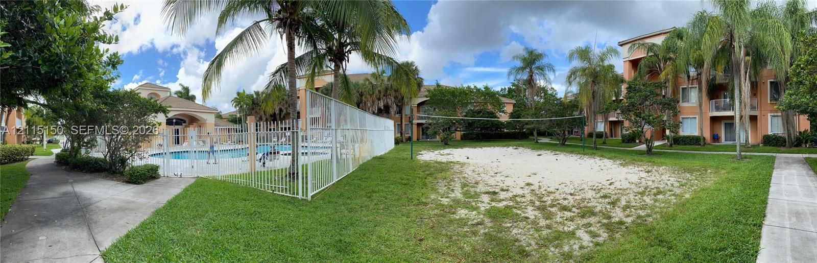 Outdoor recreation area featuring a sand volleyball court, fenced swimming pool, concrete walkways, and mature palm trees