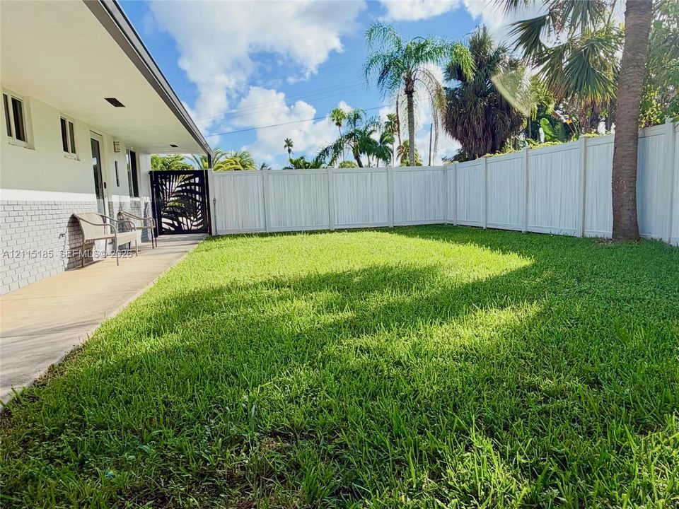 Backyard featuring a paved covered patio, manicured lawn, white vinyl fencing, and an ornate metal gate