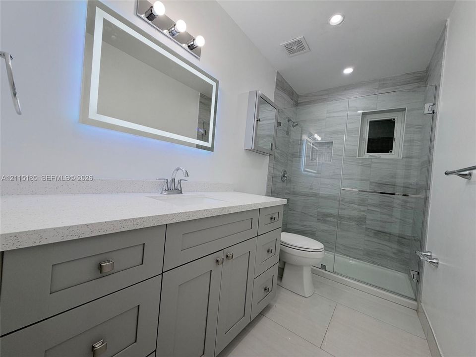Grey shaker-style vanity with a light-toned solid surface countertop, rectangular undermount sink, illuminated mirror, and glass-enclosed shower featuring horizontal-pattern wall tiling