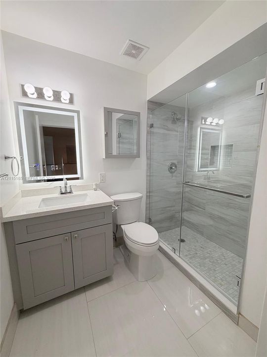 Bathroom featuring a vanity with a rectangular sink and stone-finish countertop, backlit mirror, grey cabinetry, and a glass-enclosed shower with tiled walls and flooring