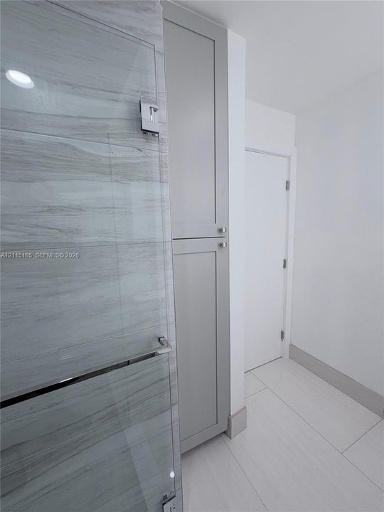 Floor-to-ceiling cabinetry with gray shaker-style doors, glass-enclosed shower featuring horizontal stone-patterned tile, and large-format porcelain tile flooring
