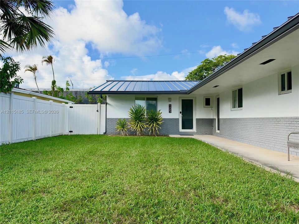 Exterior facade featuring a standing-seam metal roof, white stucco walls with grey brick lower siding, and a white vinyl privacy fence enclosing a grass lawn