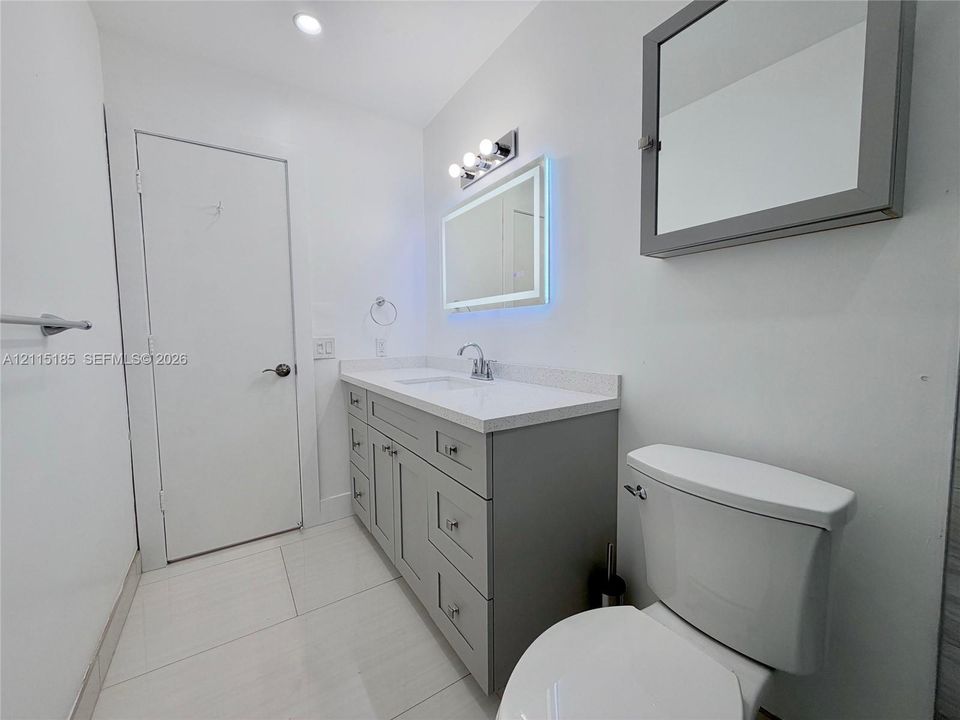 Bathroom featuring a gray vanity with a stone countertop, an integrated sink, an illuminated vanity mirror, and large-format tile flooring