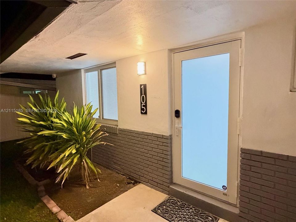 Exterior entry featuring a glass-panel door, lower brick masonry siding, a wall-mounted light fixture, and vertical address signage