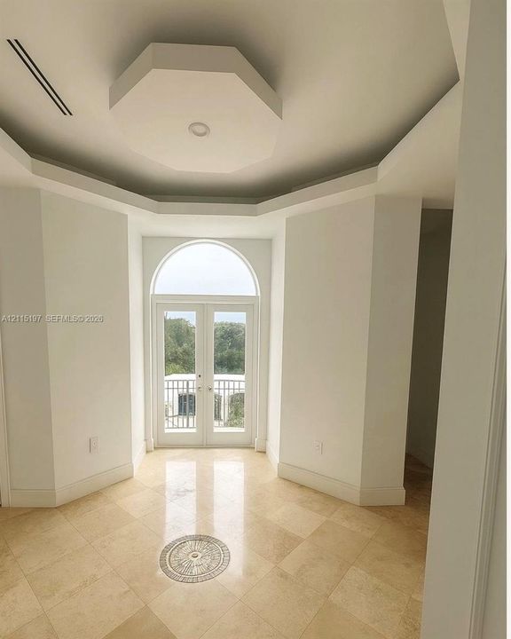 Octagonal tray ceiling, stone tile flooring with a central inlay, arched glass double doors, and a wrought-iron balcony railing