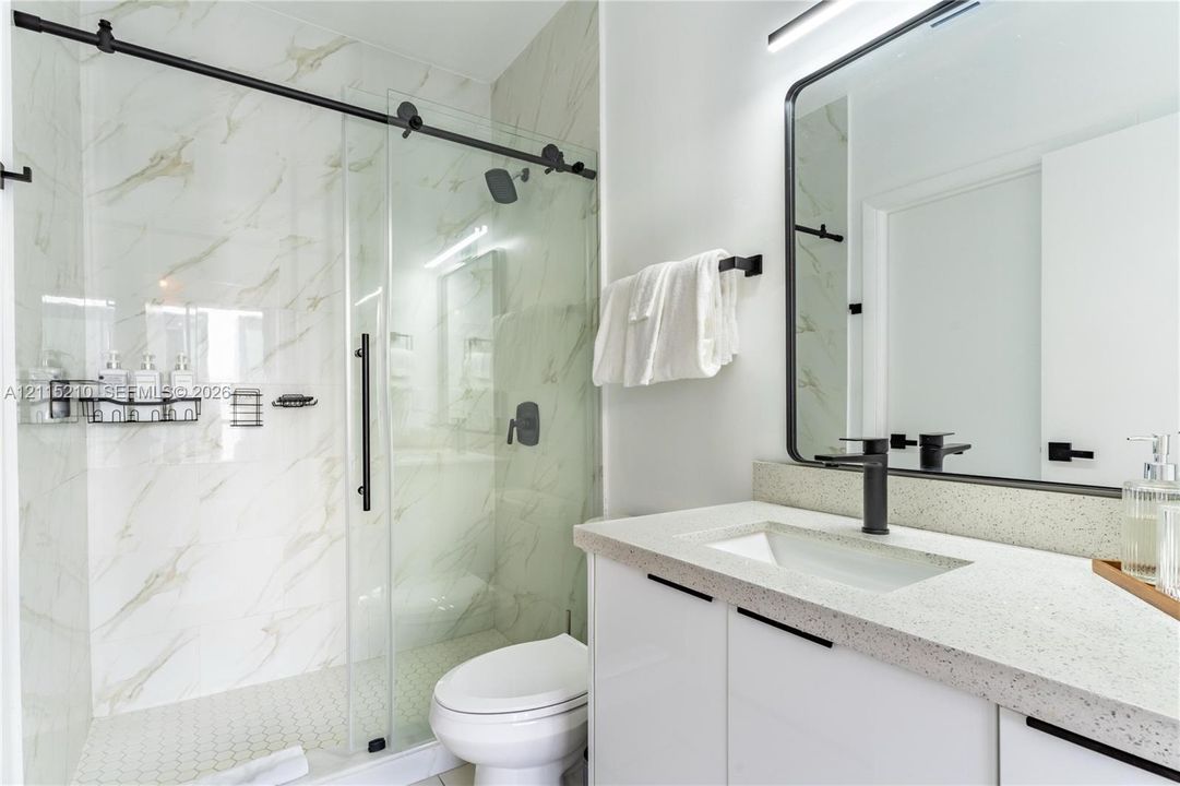 Walk-in shower with sliding glass door and stone-patterned wall tile, vanity with speckled countertop, rectangular undermount sink, matte black fixtures, and framed vanity mirror