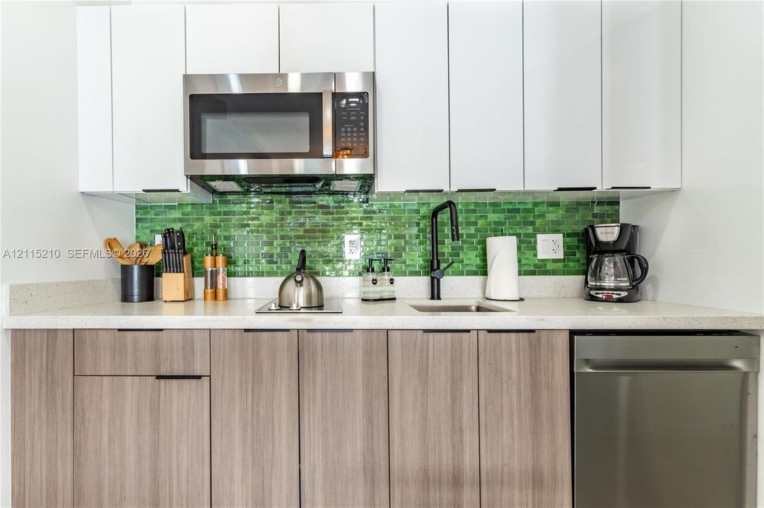 Kitchen featuring white flat-panel cabinetry, wood-finish lower cabinetry, a green glass tile backsplash, light-toned stone-finish countertops, and stainless steel appliances