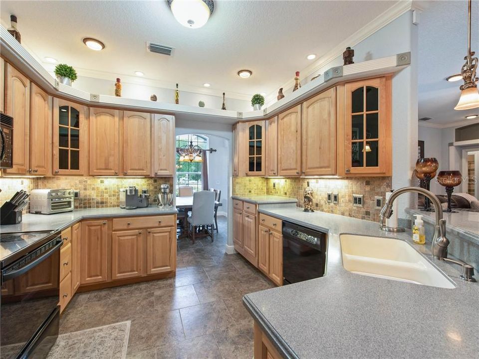 Beautiful Kitchen with staggered ceramic tile, maple raised cabinetry, double trash roll-out, lazy susan, diagonal upper cabinets with smoked glass, DuPont Corian Countertops with an Integral sink, pull-out faucet, soap dispenser, plant shelf, recess lighting with decorative trim, lovely pendant lights over the bar top, decorative tiled backsplash. View from the Butler Bar looking through the Kitchen into the Formal Dining Room.