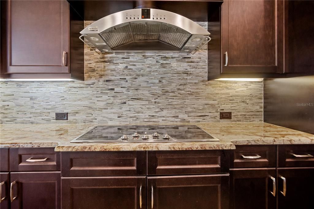 Kitchen showing details of monogram/vent and 5 burner cooktop set in granite and surrounded by tile and wooden cabinetry.