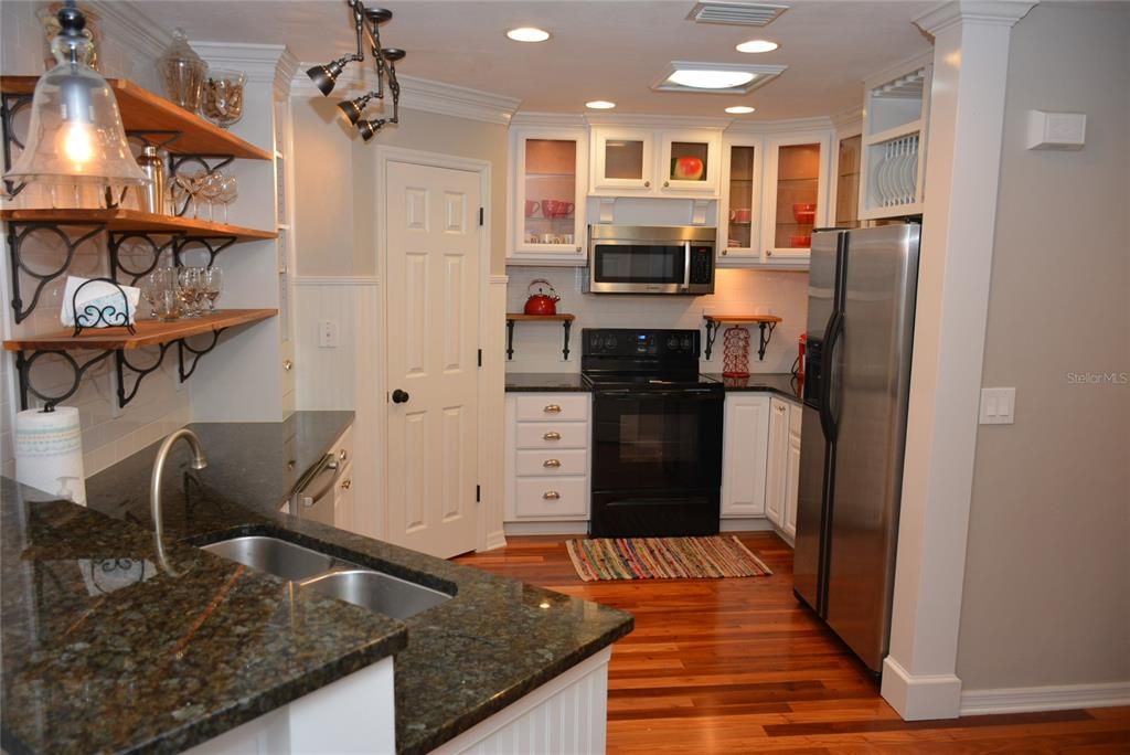 Kitchen has granite countertops, open slab wood shelving, white subway tile backsplash, crown molding, breakfront glass cabinets with in-cabinet lighting and pantry