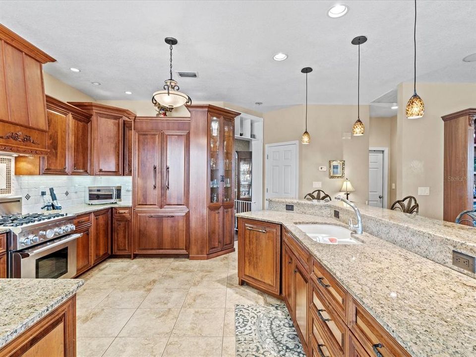 Side View of the Large, Fully Renovated Custom Kitchen w/ Custom Cherry Wood Cabinetry including Panel Fridge/Freezer, Panel Dishwasher, Custom Matching Range Hood, Gas Burner Range & Electric Oven, Undermount Sink, Glass Door China Style Cabinet, Tile Backsplash, and Passthrough Window to the Pool Area