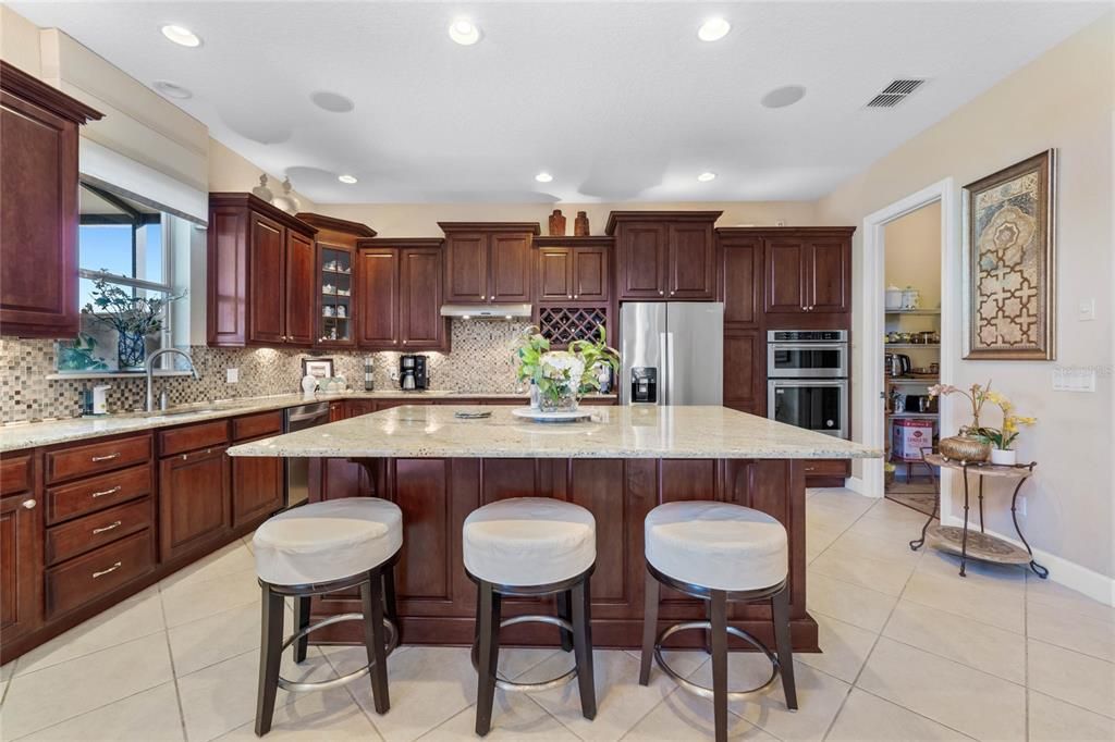 The well-appointed kitchen boasts an oversized island with counter seating, stainless appliances, granite counters, 42” solid wood cabinetry, custom tile backsplash, range hood, walk-in pantry, additional casual breakfast seating area, and a butler’s pantry